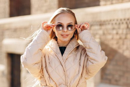 Téléchargez les photos : Portrait d'une jeune belle fille avec des cheveux lâches et des lunettes sur les yeux, portant un pull noir et un imperméable beige sur le fond d'un bâtiment en brique - en image libre de droit