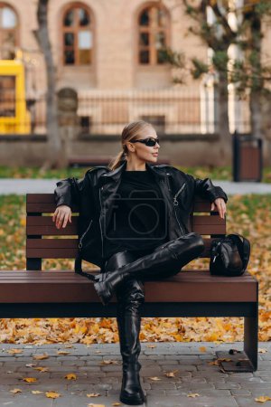 Téléchargez les photos : Une jeune femme élégante avec des lunettes de soleil noires sur les yeux regarde ailleurs, portant un t-shirt noir, une veste en cuir noir, un pantalon en cuir noir et de hautes bottes noires, tient son sac en cuir noir près et s'assoit sur un banc de parc appuyé contre une ancienne - en image libre de droit