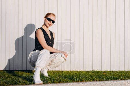Téléchargez les photos : Une femme à la mode avec des cheveux de chignon et des lunettes de soleil noires sur les yeux, portant un gilet noir, un pantalon baggy blanc et des bottes blanches à talons hauts, squats sur l'herbe près d'une clôture blanche et tient ses mains sur ses genoux - en image libre de droit