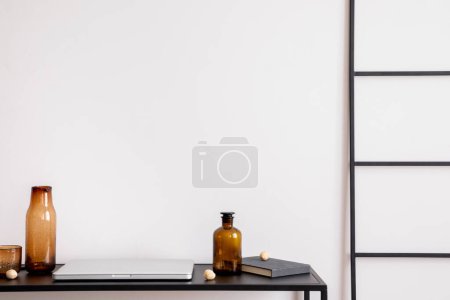Foto de Scandinavian composition with black table, glass bottle, laptop, book, dark ladder and personal accessories. Stylish and minimalist home decor. Template. - Imagen libre de derechos