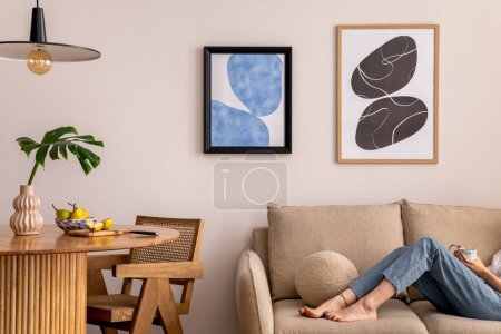 Téléchargez les photos : Warm and cozy interior of living room space with two mock up poster frames, round table, leaf in vase, chair, pedant lamp, decoration and woman lying on the sofa. Minimalist home decor. Template. - en image libre de droit