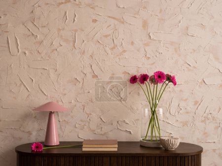 Minimalistische Wohnzimmerkomposition mit Holzanrichte, Glasvase mit rosa Blumen, Lampe, grauer Schatulle, Büchern, struktureller Wand und persönlichen Accessoires. Wohnkultur. Vorlage.