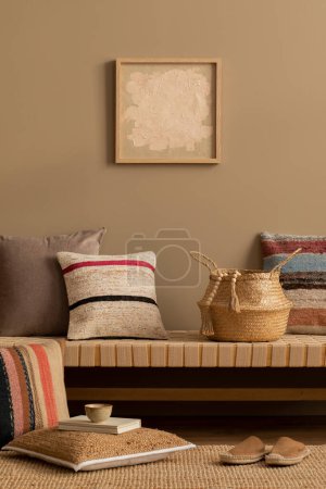 Photo for Interior design of ethno living room interior with mock up poster frame, colorful pillows, braided basket, rug, beige book, cup, brown wall, wooden floor and personal accessories. Home decor. Template - Royalty Free Image