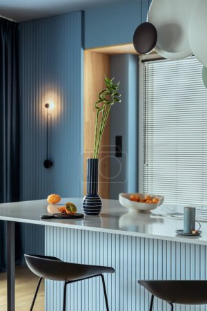 Interior design of kitchen interior with marble kitchen island, blue wall, black chokers, bowl with fruits, big window, cup, wooden floor, lamp on wall and personal accessories. Home decor. Template.