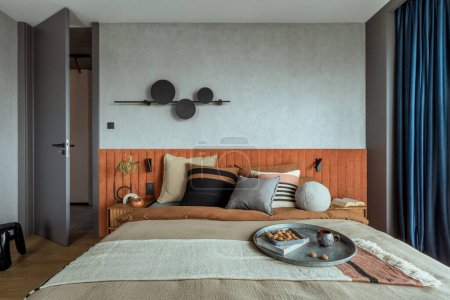 Interior design of elegant bedroom with big orange bed, beige and grey bedclothes, blue curtain, modern lamp, night stand, vase with dried flowers and personal accessories. Home decor. Template.