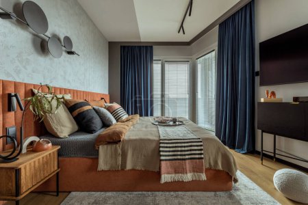 Warm and cozy bedroom interior with big orange bed, beige and grey bedclothes, blue curtain, tv, wooden night stand, black consola, round pillow, and personal accessories. Home decor. Template.