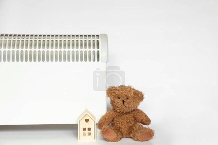 Photo for Electric convector, a small wooden house and a teddy bear next to it on a white background, a warm home - Royalty Free Image