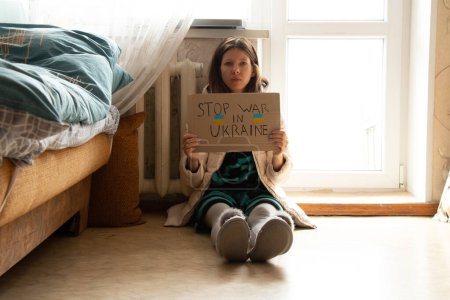 Photo for A girl sits in a winter jacket sits near a cold radiator in an apartment with a sign stop war in Ukraine, there is no heat due to the shelling of Ukraine 2022 - Royalty Free Image