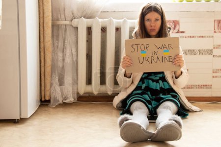 Photo for A girl sits in a winter jacket sits near a cold radiator in an apartment with a sign stop war in Ukraine, there is no heat due to the shelling of Ukraine - Royalty Free Image