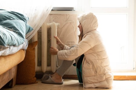 Photo for A girl in a winter jacket is sitting near a cold battery in an apartment in Ukraine, there is no heat due to shelling in Ukraine, a cold house 2022 - Royalty Free Image