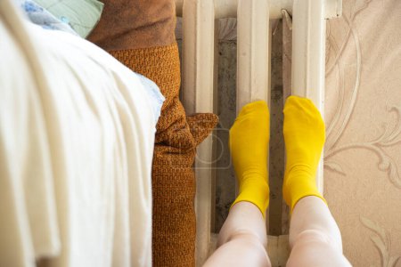 Photo for Womens legs in yellow socks are heated on a cast-iron radiator in winter in a cold apartment - Royalty Free Image