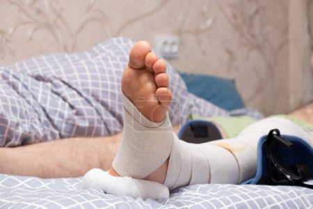 Téléchargez les photos : Jambe d'homme avec un bandage élastique et une orthèse du genou après avoir remplacé le mini à la maison au lit, la jambe après l'opération - en image libre de droit