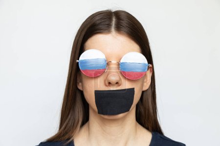 Téléchargez les photos : Drapeau de la Russie sur les lunettes d'une fille et une bouche scellée avec un bandage sur un fond blanc, la propagande sourde, aveugle et trompée de la Russie - en image libre de droit