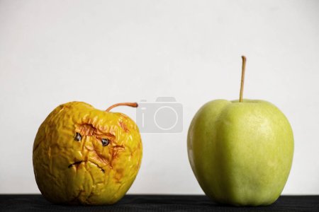 A rotten and sluggish apple with a sad painted smile and a fresh ripe green apple next to it on a white background on a black table, bad and good apples, a choice