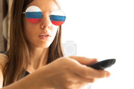Téléchargez les photos : Le drapeau de la Russie sur les lunettes de la jeune fille et dans les mains de la télécommande de la télévision, la Russie et la propagande de la télévision, mensonges et tromperie des gens par la télévision - en image libre de droit