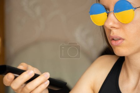 Téléchargez les photos : Le drapeau de l'Ukraine sur les lunettes d'une fille qui regarde la télévision à la maison avec une télécommande dans ses mains, un patriote - en image libre de droit
