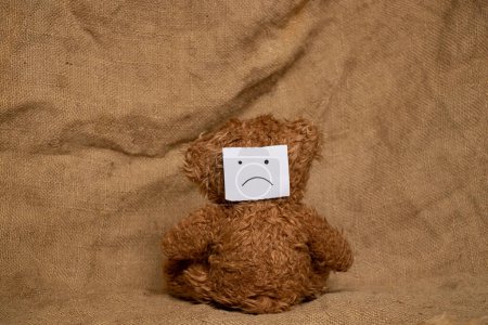 Photo for A brown bear sits with his back, and on his back there is a paper drawn with a sad face, against the background of burlap, sadness and resentment - Royalty Free Image