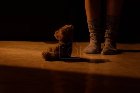 A brown children's bear sits at night on the floor of a house in an apartment and next to women's feet in socks, a children's toy