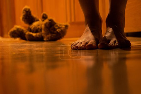 A brown children's bear sits at night on the floor of a house in an apartment and next to a woman's legs, a children's toy