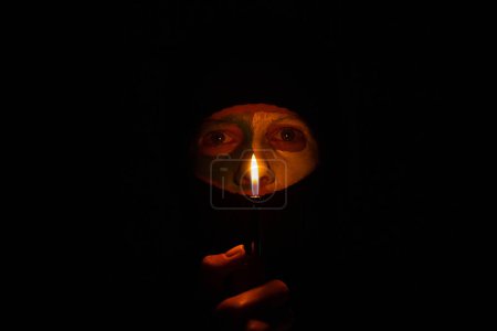 The face of a girl in a black balaclava and the flag of Ukraine are painted on her face with paints and fire from a lighter in the dark