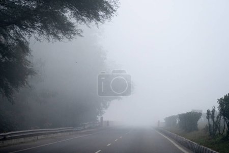 Téléchargez les photos : Early morning drives on empty road with trees bushes surrounding it with dense fog showing the cold chilly morning in Delhi, Rajasthan, Haryana India - en image libre de droit