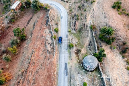 Aerial drone shot showing blue car moving on narrow country rural road with barren land on the side with green trees showing village town roads Udaipur India