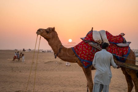 Mann im traditionellen Kurta-Pyjama-Kleid steht mit Kamel in bunten Kleidern inmitten von Sanddünen