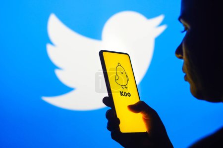 Foto de 18 de noviembre de 2022, Brasil. En esta ilustración fotográfica, la silueta de una mujer sostiene un teléfono inteligente con un logotipo de aplicación Koo que se muestra en la pantalla y el logotipo de Twitter que se muestra en el fondo - Imagen libre de derechos