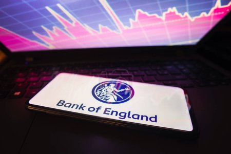 Foto de 21 de marzo de 2023, Brasil. En esta foto ilustración el logotipo del Banco de Inglaterra se muestra en una pantalla de teléfono inteligente, con la representación gráfica del mercado de valores en el fondo - Imagen libre de derechos