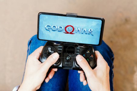 Foto de 8 de agosto de 2023, Brasil. En esta ilustración fotográfica, una persona que juega en un joystick y el logotipo de God of War se muestra en la pantalla de un teléfono inteligente - Imagen libre de derechos