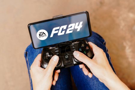 Photo for August 8, 2023, Brazil. In this photo illustration, a person playing on a joystick and the EA Sports FC 24 logo displayed on a smartphone screen - Royalty Free Image