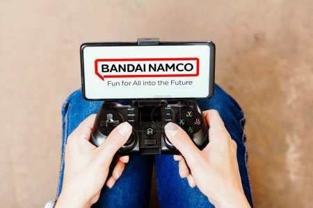 Foto de 8 de agosto de 2023, Brasil. En esta ilustración fotográfica, una persona que juega en un joystick y el logotipo de Bandai Namco Entertainment se muestran en la pantalla de un teléfono inteligente - Imagen libre de derechos