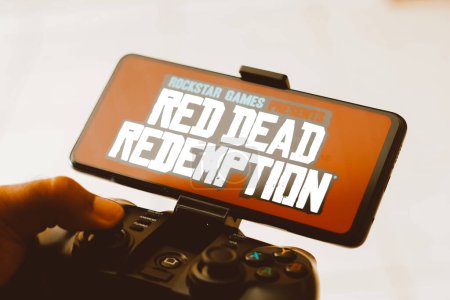Foto de 9 de agosto de 2023, Brasil. En esta ilustración de la foto, una persona que juega en un joystick y el logotipo Red Dead Redemption se muestra en la pantalla de un teléfono inteligente - Imagen libre de derechos