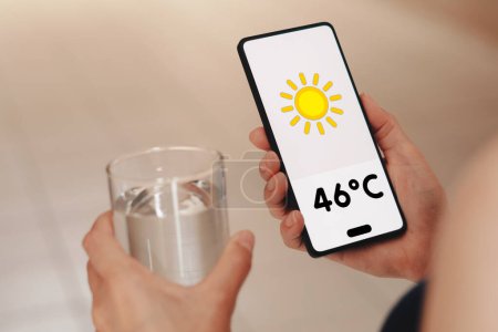 Foto de 16 de noviembre de 2023, Brasil. En esta ilustración fotográfica, una mujer sostiene un vaso de agua, el termómetro que muestra 46C de calor se muestra en la pantalla de un teléfono inteligente. - Imagen libre de derechos