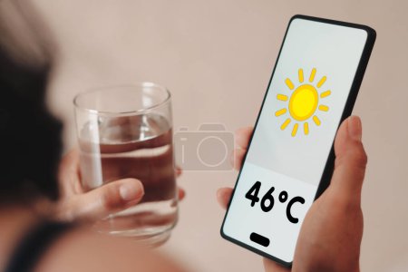 Téléchargez les photos : 16 novembre 2023, Brésil. Dans cette illustration photo, une femme tient un verre d'eau, le thermomètre montrant 46C de chaleur est affiché sur l'écran d'un smartphone. - en image libre de droit
