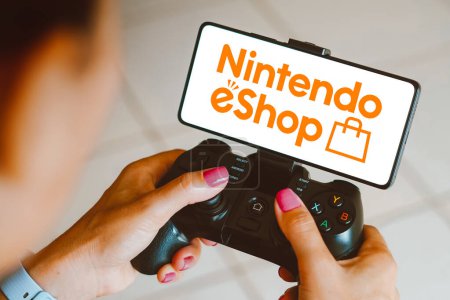 Foto de 4 de diciembre de 2023, Brasil. En esta ilustración fotográfica, el logotipo de Nintendo eShop se muestra en la pantalla de un smartphone mientras una persona juega en un mando de juego - Imagen libre de derechos