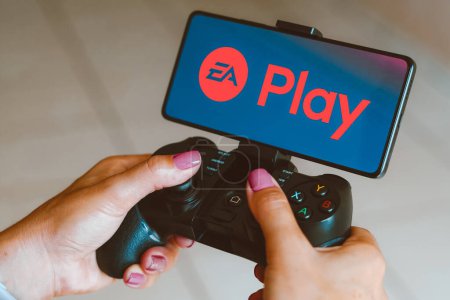 Photo for December 4, 2023, Brazil. In this photo illustration, the EA Play logo is displayed on a smartphone screen as a person plays on a gaming gamepad - Royalty Free Image