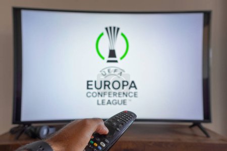 Foto de May 14, 2024, Brazil. In this photo illustration, a hand holding a TV remote control in front of the UEFA Europa Conference League (UECL) logo on a TV screen - Imagen libre de derechos
