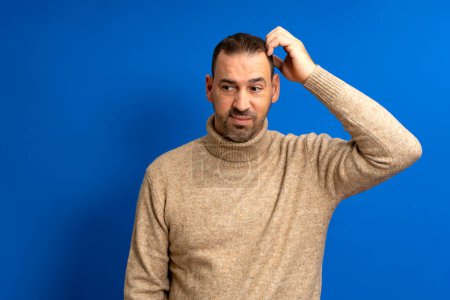 Doubtful bearded latin man thinking, scratching his head and trying to find a solution, isolated on blue background.