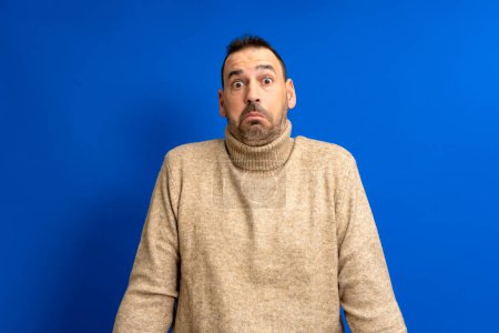 Photo for Frontal portrait of a hispanic man with a beard wearing a brown turtleneck doubting shrugging isolated on a blue background - Royalty Free Image