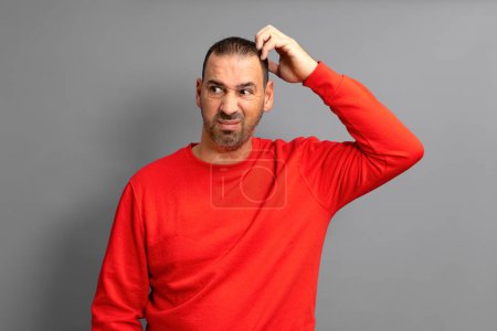 Bearded Hispanic man in red sweater frowning and looking dissatisfied while scratching his head, standing over blue background.