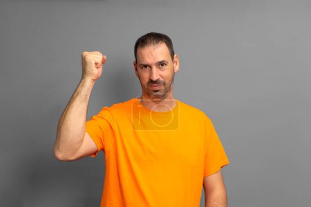 Photo for Bearded hispanic man wearing an orange t-shirt celebrating something with his fist up isolated on gray studio background - Royalty Free Image