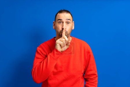 Téléchargez les photos : Serious handsome bearded hispanic man puts his index finger to his lips, tries to keep conspiracy, looks aside and says: Shh, be quiet please. Isolated shot of attractive man shows silence sign - en image libre de droit