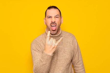 Photo for Bearded hispanic man wearing turtleneck isolated over yellow background showing rock gesture with fingers and sticking out tongue. Concept of rebellion and anarchism - Royalty Free Image