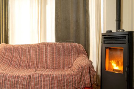 Photo for Living room with comfortable worn sofa with a cover covering it. Black cast iron pellet stove at home. Ecological heating system. Modern pellet fireplace in the interior. - Royalty Free Image