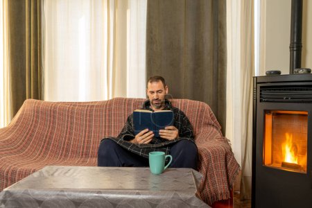 Hispanic man with a beard in his 40s wearing a bathrobe reading relaxed on the couch at home in the heat of the fire of a pellet stove