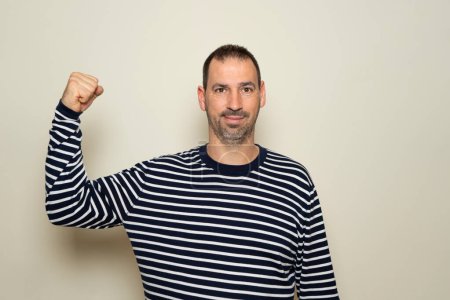 Photo for Hispanic man with a beard of 40 years old showing biceps believing that his training has paid off, the poor deluded man does not realize his terrible physical condition. Isolated beige background - Royalty Free Image