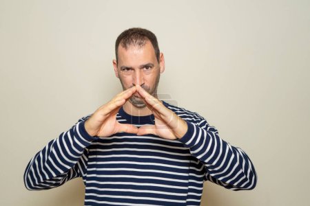Hispanic man with a beard with his fingertips together in an expression of evil, cunning and cold planning a crime. Isolated on beige background