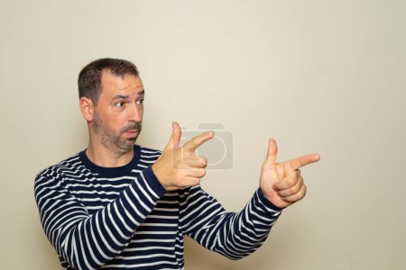 Hispanic man with beard with two hands arms sign gesture pointing aside, isolated beige wall background. Feelings, signs and facial expression symbols of positive human emotions.