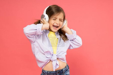 Foto de Una niña pequeña con auriculares en un fondo rosa escucha música - Imagen libre de derechos